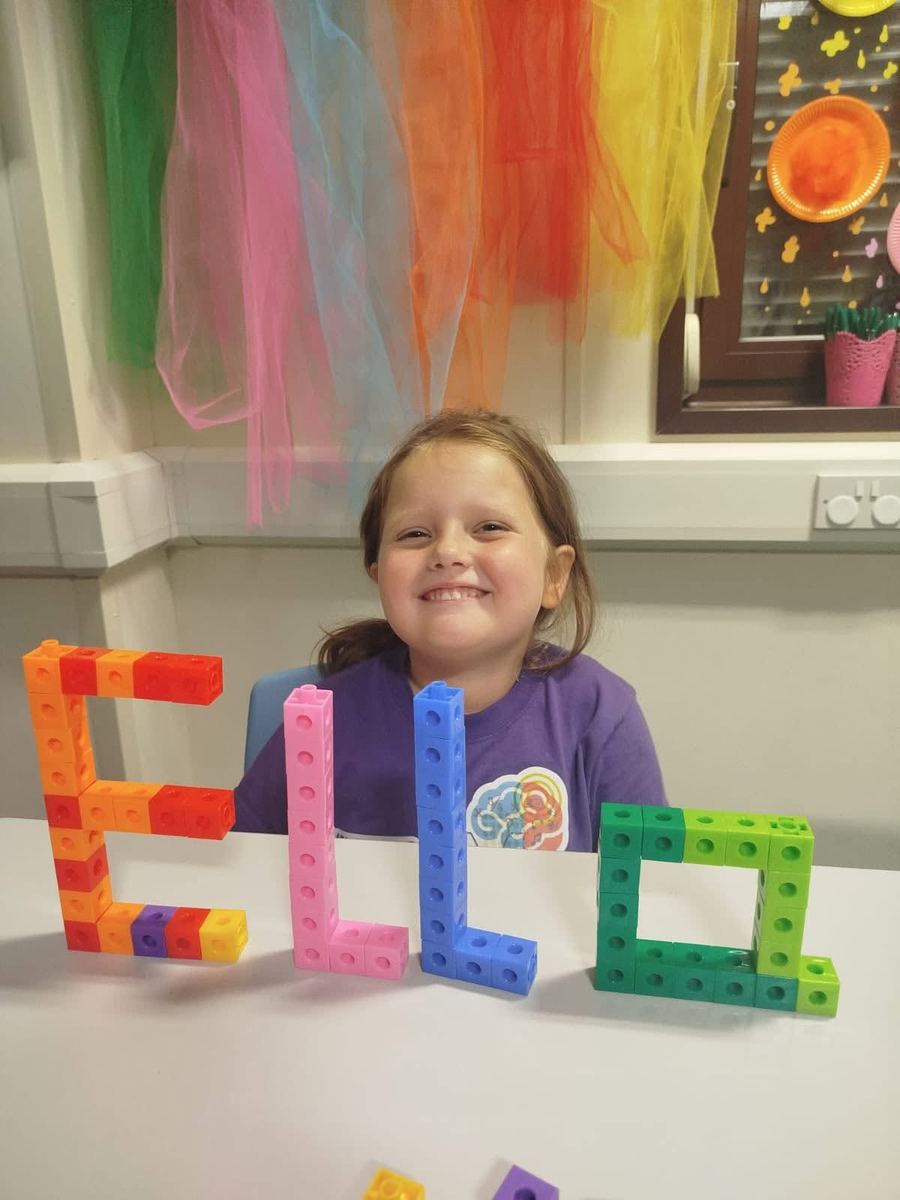A smiling learner in the classroom behind her name built from colourful blocks, with rainbow drapes behind her